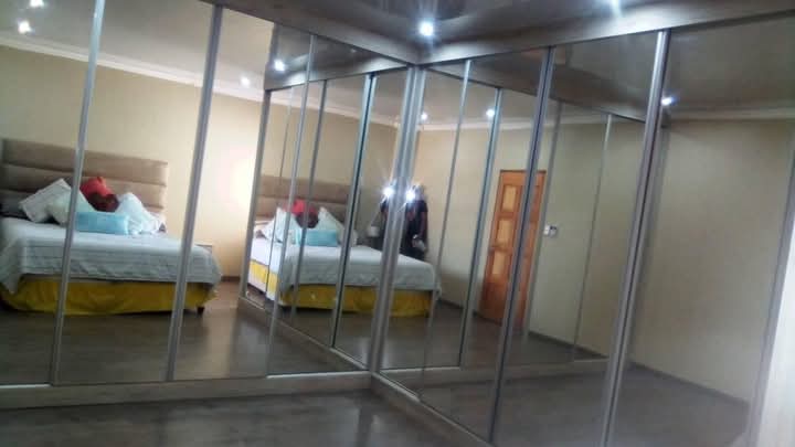 Bedroom with yellow bedding reflected in a large L-shaped mirrored sliding door wardrobe.